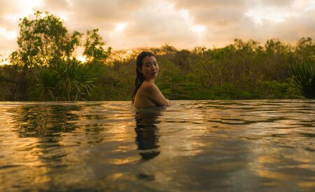 tropical holidays lifestyle portrait of young beautiful and happy Asian Japanese woman in bikini enjoying sunset at amazing jungle resort infinity pool swimming relaxed in Summer vacationの写真素材