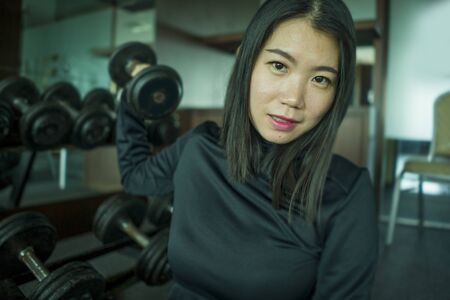 young beautiful and happy Asian Chinese woman in sport hoodie posing cool at fitness center sitting by dumbbells rack during lifting workout in body care and healthy lifestyle conceptの写真素材