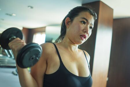 mirror portrait of young pretty and fit Asian Korean woman working hard at hotel gym or fitness center lifting dumbbell doing body building indoors workout in healthy lifestyle conceptの写真素材