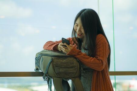 young beautiful and happy Asian Japanese woman checking mobile phone at airport departure lounge carrying backpack waiting for boarding in aircraft ready for holiday travel smiling cheerfulの写真素材