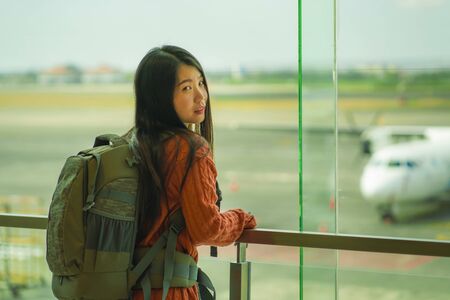 young happy and excited Asian Korean student woman with backpack at airport departure lounge watching aircraft through glass window smiling ready for boarding in holidays travelの写真素材