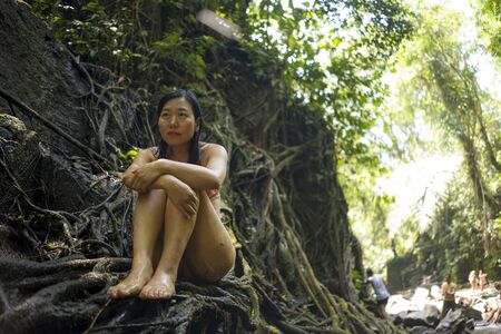 beautiful woman at tropical enchanted forest . Young attractive Asian Chinese girl in bikini sitting on tree trunk at tropical paradise jungle with a mysterious and magical feeling in wanderlust conceptの写真素材