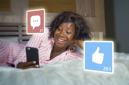 young beautiful and happy black afro American woman in pajamas using mobile phone social media...