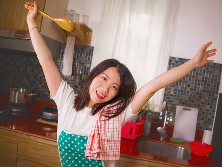 natural lifestyle portrait of young beautiful and happy Asian Chinese woman in kitchen apron holding spoon smiling cheerful enjoying domestic chores and housekeeping posing playful and sweetの写真素材