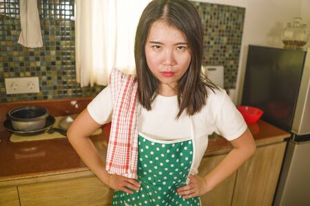 domestic chores lifestyle portrait of young tired and stressed Asian Korean woman in cook apron working in the kitchen looking angry and moody feeling unhappy and overworked at homeの写真素材