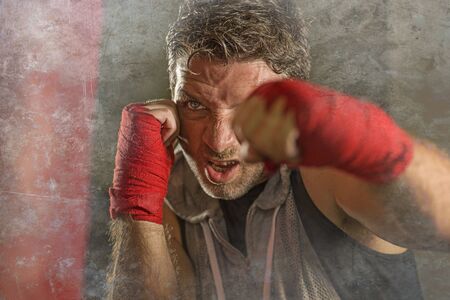 lifestyle gym portrait of young attractive and fierce looking man training boxing at fitness club doing heavy bag punching workout with hands and wrist wraps in badass fighter lookの写真素材