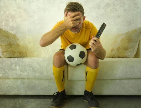funny portrait of young stressed and excited football fan man watching soccer game on TV at home couch dressed in team player uniform feeling the stress and emotion of the competitionの写真素材