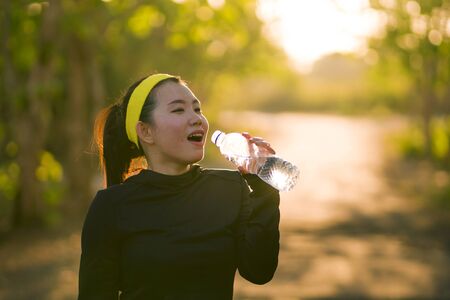 young Asian runner girl drinking water sweaty and tired. Attractive and happy Korean woman thirsty after running workout and jogging at beautiful park on sunset in fitness lifestyleの写真素材
