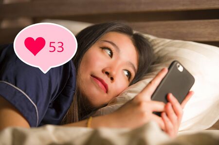 Young beautiful and happy Asian girl getting social media likes. Gorgeous Chinese woman in bed at...