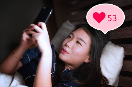 Young beautiful and happy Asian girl getting social media likes. Gorgeous Chinese woman in bed at night excited getting positive feedback on internet  app smiling cheerfulの写真素材