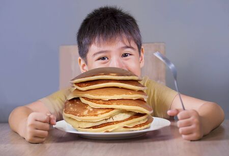 Young boy having pancakes breakfast. 7 or 8 years old happy and excited child sitting on table eating huge pile of pancakes smiling naughty in sugar abuse and unhealthy nutritionの写真素材