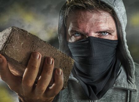 young man as ultra and radical anarchist rioter . furious antifa protester in face mask throwing brick hostile on street on demonstration in violet riot and chaosの写真素材