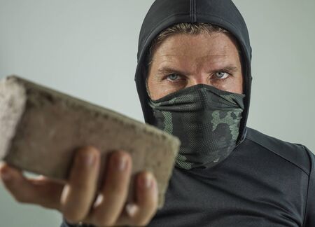 young man as radical and aggressive anarchist rioter holding brick threatening. furious anti-system protester in face mask throwing stone in violent riot  isolated on white backgroundの写真素材