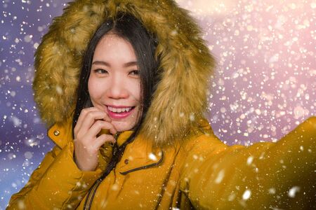 young beautiful and happy Asian Korean woman in hood winter jacket taking selfie portrait with mobile phone outdoors under the snow smiling cheerful enjoying Christmas holidays tripの写真素材