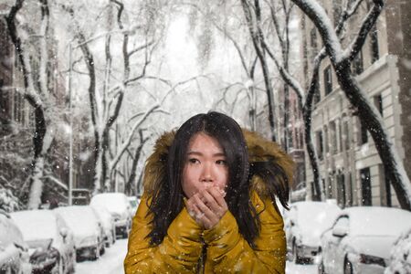 young beautiful Asian Korean woman feeling cold and chilly freezing feeling cold in Winter weather wearing yellow jacket with fur hood rubbing her hands to get warm under snowfallの写真素材
