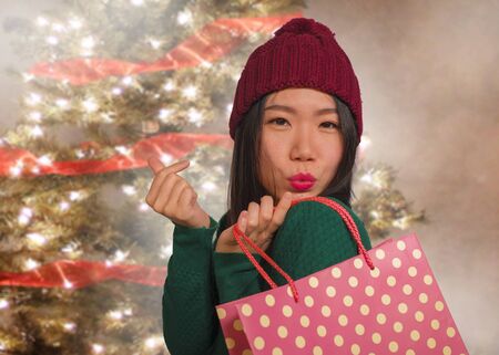 lifestyle portrait of young happy and beautiful Asian Korean woman in Winter hat holding shopping bag Christmas present excited and cheerful isolated on X-mas decoration backgroundの写真素材
