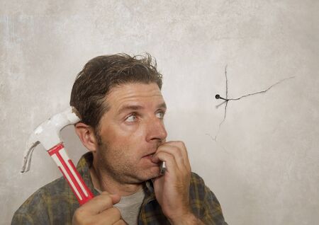 funny portrait of man holding hammer after driving a nail for hanging a frame but making a mess cracking the wall as a disaster DIY guy and messy domestic repair task の写真素材