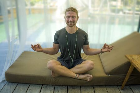 hipster guy practicing yoga . young attractive and happy athletic man sitting on resort bed in lotus pose doing meditation exercise smiling relaxed and zen like in harmony and inspiration conceptの写真素材
