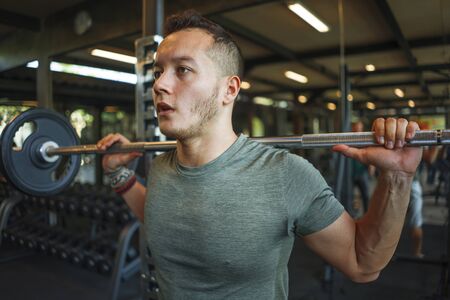 indoors fitness portrait of young athletic and sweaty mixed ethnicity man doing squats with barbell...