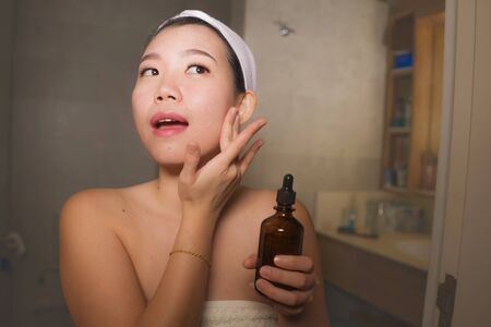 natural lifestyle portrait of young beautiful and happy Asian Chinese woman applying anti aging serum cosmetic on her face at home bathroom smiling fresh in facial skin care and beauty conceptの写真素材