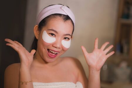 young beautiful and happy Asian Chinese woman smiling to bathroom mirror applying hydrating eye patches under the eyes to repair dark circles in beauty cosmetics and facial skin care conceptの写真素材