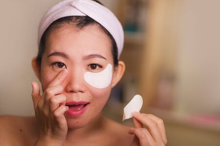 young beautiful and happy Asian Japanese woman smiling to bathroom mirror applying hydrating eye patches under the eyes to repair dark circles in beauty cosmetics and facial skin care conceptの写真素材