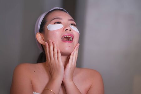 young beautiful and happy Asian Korean woman smiling to bathroom mirror applying hydrating eye patches under the eyes to repair dark circles in beauty cosmetics and facial skin care conceptの写真素材