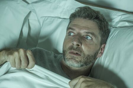 dramatic portrait of stressed and scared man alone in bed awake at night in fear after having a nightmare feeling paranoid holding the blanket in funny panic face expressionの写真素材
