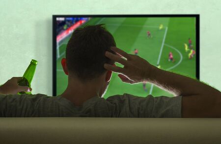 view from behind of young soccer fan man watching European football game on big screen television at...