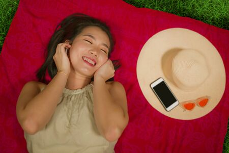 outdoors lifestyle portrait of young happy and beautiful Asian Chinese student girl chilling on city park relaxed listening to music with earpiece lying flat on sarong over green grass delightedの写真素材