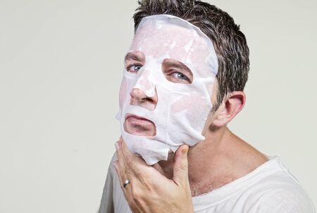 lifestyle isolated background portrait of young weird and funny man at home trying using paper facial mask cleansing applying anti aging beauty treatment in hilarious face expressionの写真素材