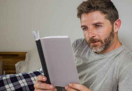 lifestyle portrait of young attractive and relaxed man at home lying comfortable on bed wearing  pajamas reading a book enjoying an interesting novel chilled and cozyの写真素材