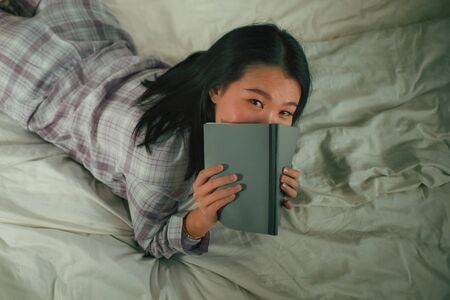 lifestyle portrait of young beautiful and sweet Asian Korean woman in bed reading book novel feeling happy and playful or studying at home wearing cute pajamas looking cozy and homeyの写真素材