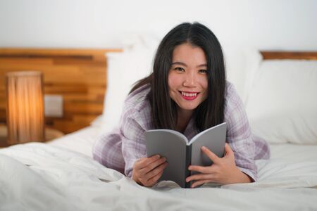 lifestyle portrait of young beautiful and sweet Asian Korean woman in bed reading book novel feeling happy and playful or studying at home wearing cute pajamas looking cozy and homeyの写真素材