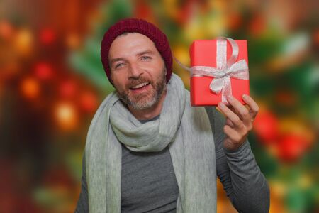 lifestyle portrait of young happy and attractive man in winter hat and scarf smiling holding Christmas gift box receiving or giving present isolated on xmas tree lights bokeh backgroundの写真素材