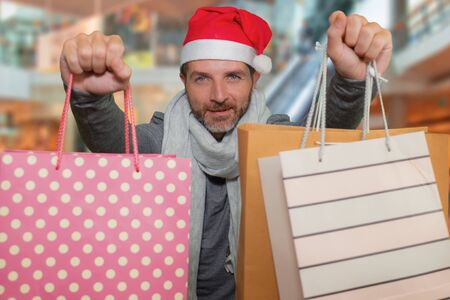lifestyle portrait of young happy and attractive hipster man in Santa hat and scarf smiling holding lot of shopping bags buying Christmas presents excited and cheerful at shopping mallの写真素材