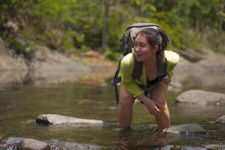 young beautiful and happy Asian Chinese woman carrying backpack trekking on mountains crossing river...