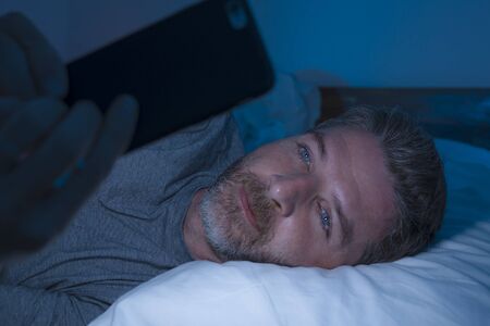 young attractive and happy man with blue eyes lying on bed late at night in dark light networking on mobile phone or online dating smiling relaxed enjoying internet in social media addiction conceptの写真素材
