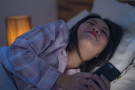 bedtime lifestyle portrait of young beautiful and happy sweet Asian Chinese woman with in pajamas enjoying with mobile phone app in bed at night in internet social media addiction conceptの写真素材