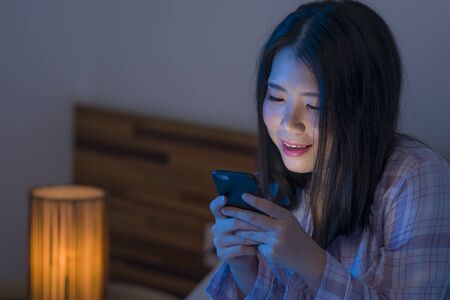bedtime lifestyle portrait of young beautiful and happy sweet Asian Korean woman with in pajamas enjoying with mobile phone app in bed at night in internet social media addiction conceptの写真素材