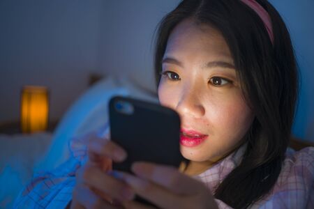internet addict Asian girl mesmerized with hand phone , young beautiful and excited Chinese woman in pajamas enjoying with mobile phone in bed at night having fun online in social media addictionの写真素材