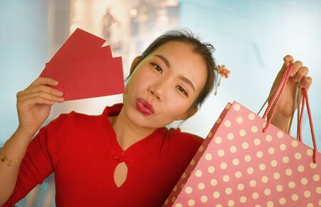lifestyle portrait of young beautiful and happy Asian Chinese woman holding Chinese New year red pockets and shopping bags at modern shopping mall smiling excited spending money in clothingの写真素材