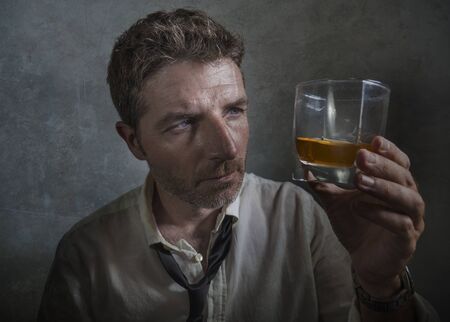 dramatic portrait of 30s to 40s alcoholic  man in lose necktie drinking alcohol desperate and wasted looking at whiskey glass thoughtful drunk and depressed fighting his drinking addictionの写真素材
