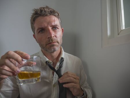 dramatic portrait of 30s to 40s alcoholic  man in lose necktie drinking desperate holding whiskey glass thoughtful drunk and depressed completely wasted in alcohol addiction conceptの写真素材