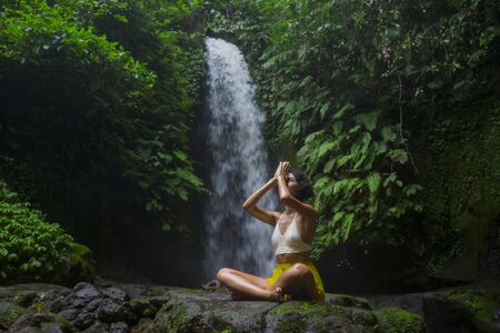 outdoors portrait of young attractive and happy hipster girl doing yoga at beautiful tropical waterfall meditating enjoying freedom and nature in healthy and zen lifestyleの写真素材