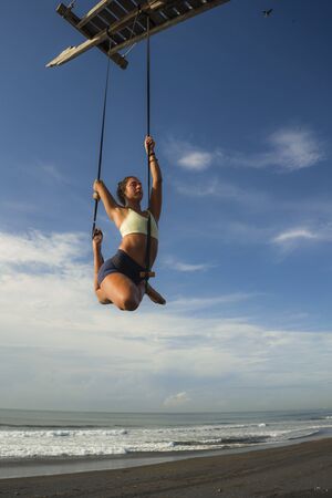 aerial yoga beach workout - young attractive and healthy woman practicing aero-yoga training balance...