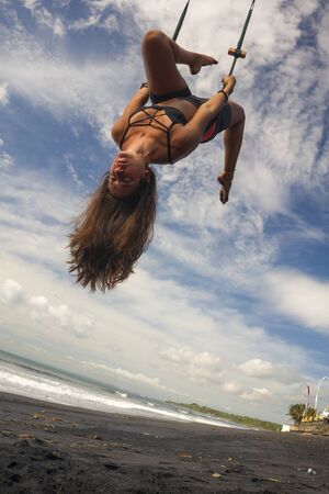 aerial yoga beach workout - young attractive and healthy woman practicing aero-yoga training balance body and mind control hanging from ropes above the sea isolated on beautiful blue skyの写真素材