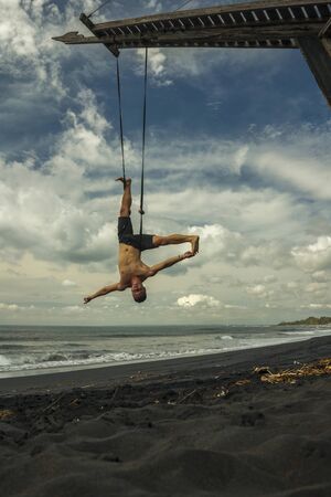 outdoors portrait of young attractive and athletic man practicing aero yoga workoutat the beach...