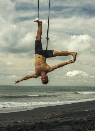 outdoors portrait of young attractive and athletic man practicing aero yoga workoutat the beach hanging from rope swiing above the sea training body balance and flexibility in healthy lifestyleの写真素材
