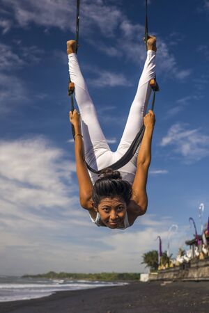 outdoors portrait of young happy and athletic Asian Indonesian woman doing aero yoga beach workout training body balance and relaxation hanging from swing rope above the sea in healthy lifestyleの写真素材